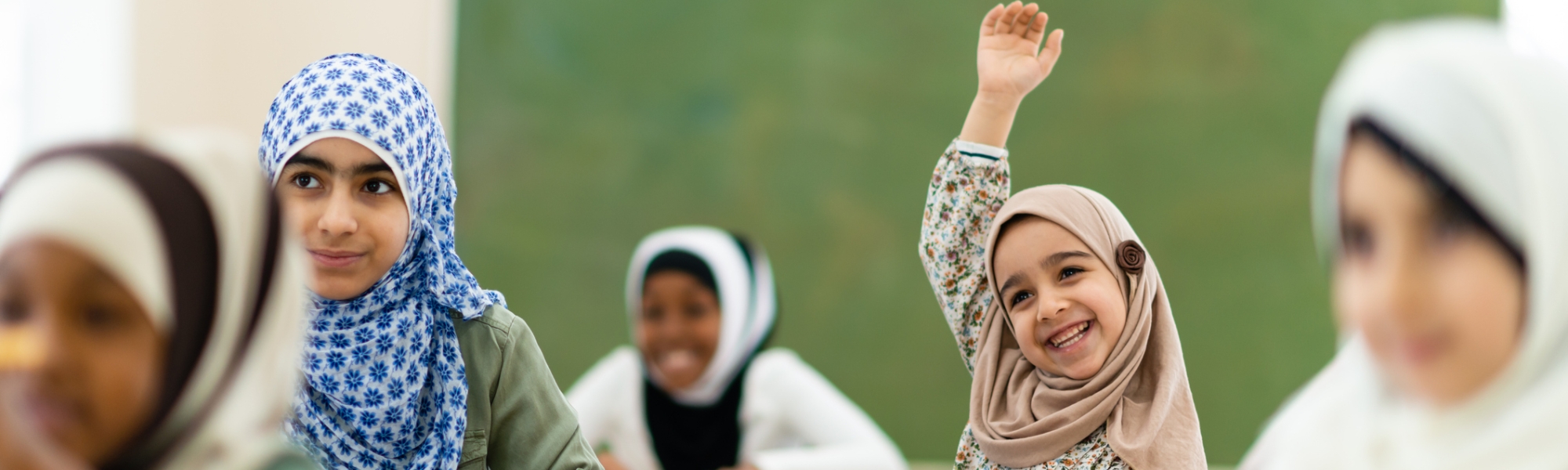 A smiling girl raising her hand to answer a question among middle school classmates wearing hijabs