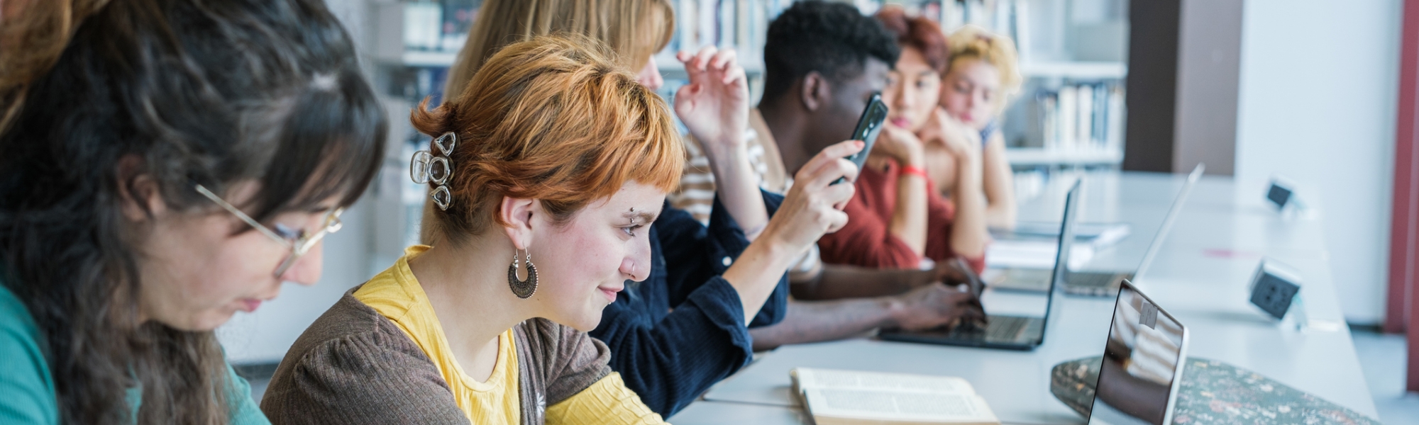 Six international students studying in the college library with books, paper, phones, and laptops