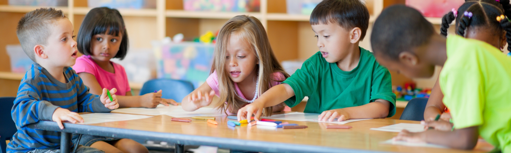 Six young children writing on paper and observing each other at a table in a classroom