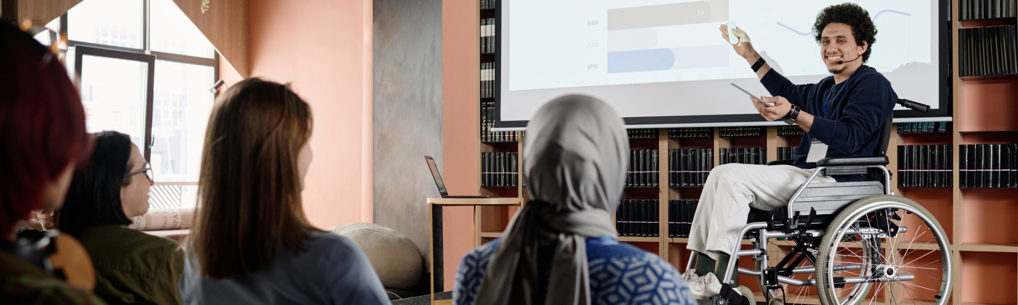 A man in a wheelchair gesturing to a slide on a screen during a graduate school presentation